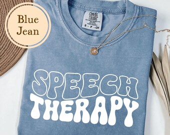 May include: A blue jean colored t-shirt with the words "SPEECH THERAPY" in white, retro-style lettering. The shirt has a crew neck and short sleeves. A gold necklace with a pendant is draped across the shirt. The shirt is made by Comfort Colors.