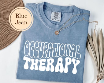 May include: Blue jean colored t-shirt with the words "OCCUPATIONAL THERAPY" in white, retro-style font. A gold necklace with a pendant rests on the shirt. The shirt is folded, with a woven mat and dried plant in the background.