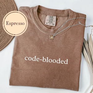 May include: Espresso-colored t-shirt with the text "code-blooded" in white. The shirt has a crew neck and a small tag at the neckline. A silver necklace with a pendant rests on the shirt.