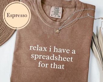 May include: Espresso-colored t-shirt with the text "relax i have a spreadsheet for that" in white. A silver necklace with a pendant rests on the shirt. The shirt is a Comfort Colors brand.