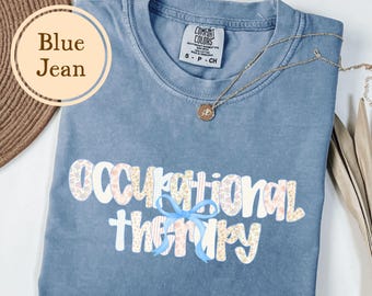 May include: Blue jean colored t-shirt with the words "Occupational Therapy" in a floral design. The shirt has a light blue bow and a gold necklace. The shirt is made by Comfort Colors.