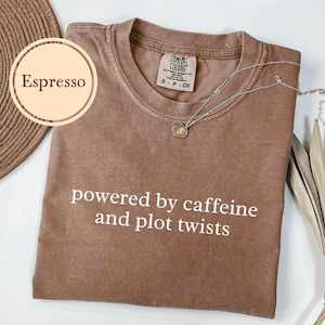 May include: Espresso-colored t-shirt with the text "powered by caffeine and plot twists" in white. A silver necklace with a small pendant rests on the shirt. The shirt is a Comfort Colors brand.