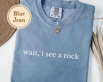 May include: A blue jean colored t-shirt with the text "wait, i see a rock" in white. The shirt has a crew neck and short sleeves. A gold necklace with a pendant is draped across the shirt.