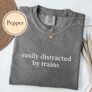 May include: A gray Comfort Colors t-shirt with the text "easily distracted by trains" in white. A gold necklace with a pendant rests on the shirt. The shirt is folded and displayed on a white surface.