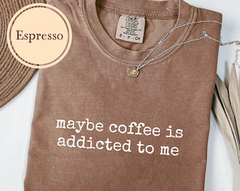 May include: Espresso-colored t-shirt with the phrase "maybe coffee is addicted to me" in white text. A silver necklace with a pendant rests on the shirt. The shirt is a Comfort Colors brand.