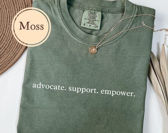 May include: A moss green t-shirt with the words "advocate. support. empower." printed in white. A gold-colored necklace with a pendant rests on the shirt. The shirt is folded and has a tag that says "Comfort Colors".