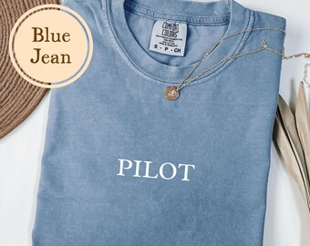 May include: A blue jean colored t-shirt with the word "PILOT" printed in white. A gold necklace with a pendant is draped across the shirt. The shirt is folded on a white surface.
