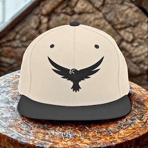 May include: A beige and black baseball cap featuring a black graphic of a bird with outstretched wings. The cap has a flat brim and a black button on top. The bird graphic is centered on the front of the cap.