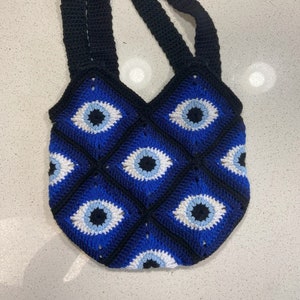 Puede incluir: Una bolsa de mano de crochet con asas negras y un diseño de ojo turco azul, blanco y negro.