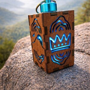 May include: A wooden lantern with a blue light shining through cut-out designs. The lantern has a blue top and a gold-colored ring. The designs include a crown and other abstract shapes. The lantern is sitting on a rock.