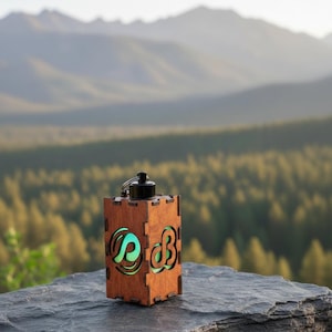May include: A wooden lantern with a black top and a green glowing interior. The lantern has cut-out designs on the sides. The background features a scenic view of a mountain range and a forest.