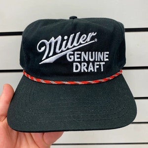 Op de afbeelding: Een zwarte baseballpet met een wit geborduurd Miller Genuine Draft logo. De pet heeft een rood en wit touwbandje rond de rand.