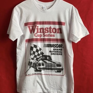 May include: White t-shirt with a red and white Winston Cup Series logo and a black and white illustration of a race car with a checkered flag. The text "NASCAR Grand National Drivers" is printed below the car.