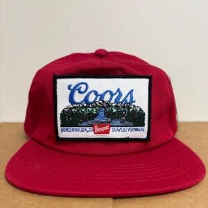Op de afbeelding: Rode baseballcap met een platte klep. De voorkant heeft een rechthoekige patch met het woord "Coors" in blauw script boven een berg- en rivierlandschap. De patch bevat ook de tekst "Brewed in Golden, CO Banquet Served Everywhere."
