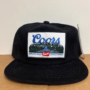 May include: Black corduroy baseball cap with a rectangular patch featuring the Coors logo. The patch displays a mountain scene with blue text and a red and white banner. The cap has a flat brim and a tag.