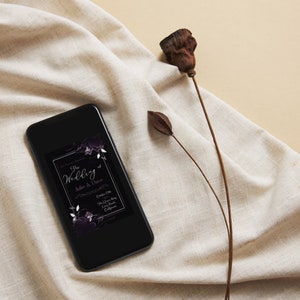 May include: A black smartphone displays a wedding invitation with floral accents. The invitation text reads "The Wedding of Julie & Dave" with event details. The phone rests on a beige fabric with a dried flower stem.