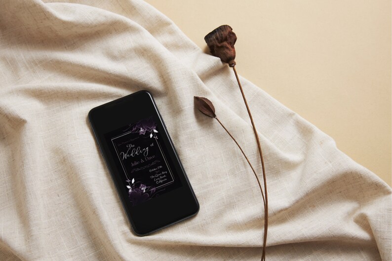 May include: A black smartphone displays a wedding invitation with floral accents. The invitation text reads "The Wedding of Julie & Dave" with event details. The phone rests on a beige fabric with a dried flower stem.