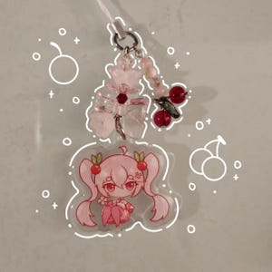May include: A pink anime-style charm featuring a character with cherry-themed hair and accessories. The charm is adorned with a butterfly-shaped embellishment, red cherry beads, and pink beads. A white cord is attached for hanging.
