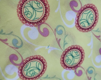 May include: Close-up of a fabric with a repeating floral pattern. The design features oval medallions with a red border and green swirls, set against a pale yellow background. White and purple floral elements complete the design.