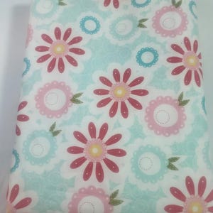 May include: A folded fabric featuring a floral pattern. The design includes large pink and red flowers with yellow centers, surrounded by light blue and pink accents on a white background. Green leaves are interspersed throughout the design.