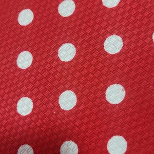 May include: Close-up of a red fabric with a woven texture, featuring a pattern of evenly spaced white polka dots. The dots are circular and appear slightly textured against the red background.