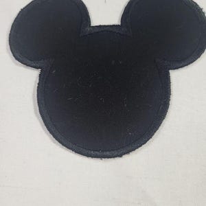May include: A solid black fabric patch shaped like a mouse head silhouette with two rounded ears and a circular face. The patch features a thick, stitched border around the entire perimeter, creating a defined edge for this iconic character shape.