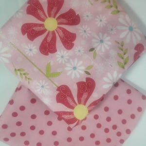May include: Two pink fabric squares. One features a large red flower with a yellow center and white daisies on a pink background. The other has a pink polka dot pattern.