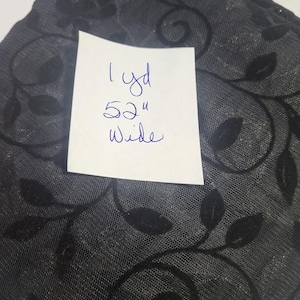 May include: Black sheer fabric featuring a velvet-like floral and vine pattern. A white paper note is placed on top with the handwritten text 1 yd 52" wide, indicating the length and width of the material in inches.
