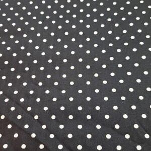 May include: Black fabric with a repeating pattern of white polka dots. The dots are evenly spaced and vary slightly in size. The fabric has a slight sheen and appears to be a medium-weight material, suitable for various sewing projects.