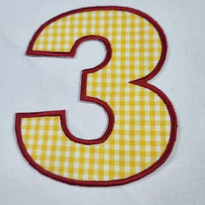 May include: A fabric applique in the shape of the number three, featuring a yellow and white gingham check pattern. The numeral is outlined with a thick, dark red embroidered border that defines its shape against the white fabric background.
