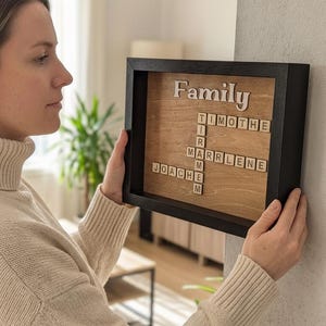 May include: A framed wooden sign with the word "Family" in a cursive font. The sign features a crossword-style arrangement of family member names in wooden letter tiles. The frame is black, and the background is a light wood.