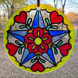 May include: A round, stained glass suncatcher with a central blue star design. The star is surrounded by a red flower and heart shapes, with green leaves. The outer edge is yellow with a scalloped design, and a red ribbon is attached for hanging.
