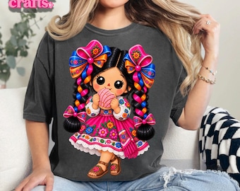Peut inclure: Un t-shirt gris anthracite avec une illustration graphique d'une poupée aux cheveux noirs tressés, portant de grands nœuds et une robe rose, tenant un pain concha rose. La poupée porte des sandales et des broderies florales sur les manches.