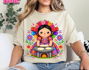 Peut inclure: Un t-shirt beige avec un motif coloré représentant une poupée couronnée de fleurs, vêtue d'une robe à motifs, utilisant un rouleau à pâtisserie sur une petite table avec un feu en dessous. Le texte Yohana crafts et Transferencia DTF Textil est visible.