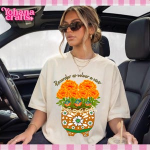 Peut inclure: Un t-shirt à manches courtes de couleur crème avec un motif de fleurs souci orange dans un pot marron à motifs. Le texte "Recordar es volver a vivir" est imprimé dans une police cursive sombre au-dessus de l'illustration florale.