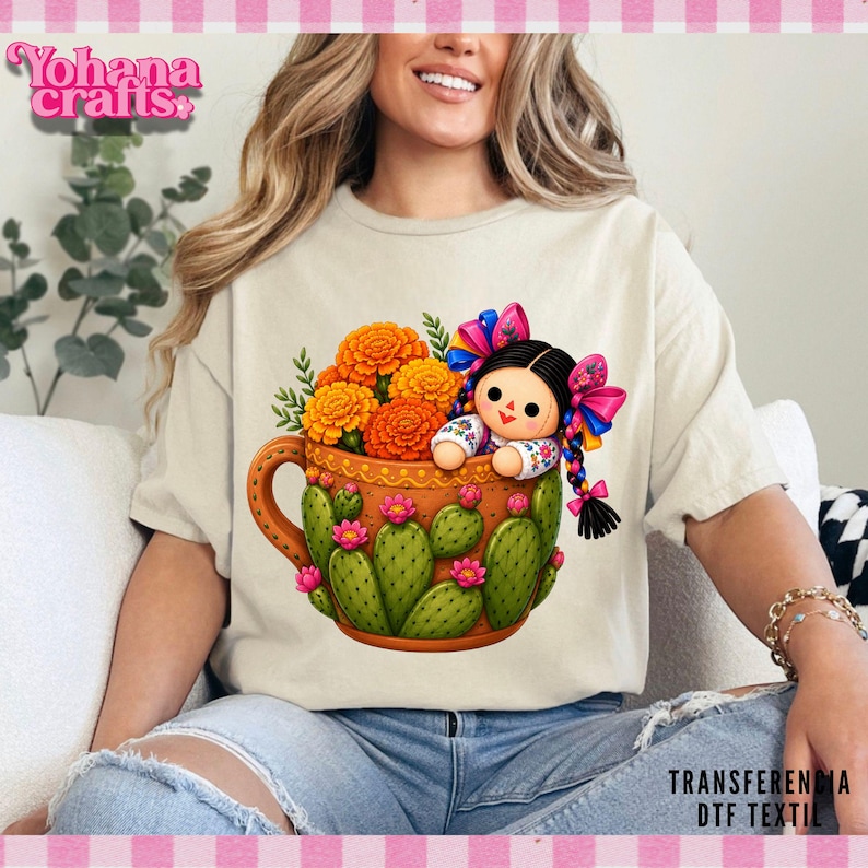 Peut inclure: Un t-shirt couleur cr&egrave;me arborant un motif de tasse marron d&eacute;cor&eacute;e de cactus verts et de fleurs roses. Dans la tasse se trouvent des soucis orange et une petite poup&eacute;e aux cheveux noirs, rubans color&eacute;s et chemise blanche brod&eacute;e.