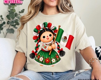 Peut inclure: Un t-shirt beige avec un motif de poupée aux cheveux tressés ornés de rubans rouges, blancs et verts. La poupée porte une robe verte à motifs floraux et tient un drapeau mexicain. Le texte "Yohana crafts" et "TRANSFERENCIA DTF TEXTIL" est visible.