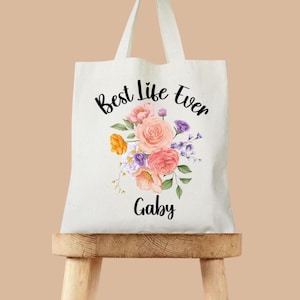 Peut inclure: Un sac fourre-tout crème avec le texte "Best Life Ever" au-dessus d'un arrangement floral de fleurs roses, oranges et violettes. Le nom "Gaby" est écrit en dessous. Le sac a des poignées blanches.