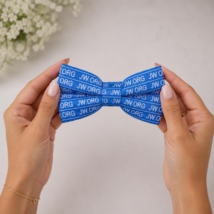 May include: A blue bow tie featuring a repeating white horizontal stripe pattern with the text JW.ORG printed in white across the fabric. The bow tie has a classic shape with a central knot and two symmetrical wings extending to the sides.