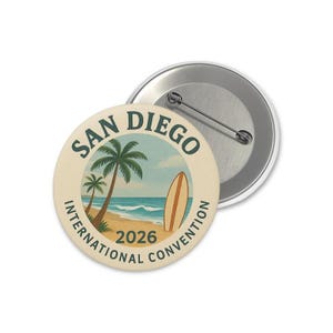 Puede incluir: Un botón redondo con una escena de playa que presenta palmeras, una tabla de surf y el texto "San Diego International Convention 2026". El botón tiene una parte trasera de metal con un alfiler.