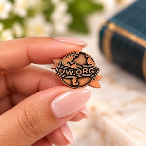 Puede incluir: Un broche circular de metal color cobre que presenta un mapa estilizado del mundo con una cinta horizontal en el centro que muestra el texto JW.ORG en letras negras negritas. El diseño tiene un acabado oscuro y profundo.