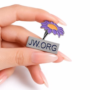 Puede incluir: Un pin de solapa de metal con una base rectangular plateada que presenta el texto JW.ORG en letras azul oscuro. En la parte superior del rectángulo hay una flor estilizada con pétalos morados y un centro amarillo, hecha de esmalte y metal.