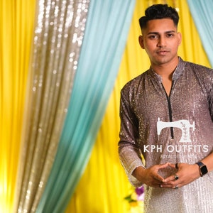 Può includere: Un uomo indossa una kurta di paillettes argentate con una cerniera nera e il testo "KPH OUTFITS ROYAL STYLE" sul davanti. È in piedi davanti a uno sfondo giallo e blu.