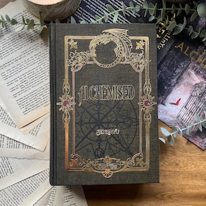 May include: A dark green hardcover book titled "ALCHEMISED" with gold foil detailing, including a dragon and floral patterns. The book is set amongst scattered pages, a lit candle, and greenery, creating a mystical and atmospheric scene.