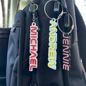 May include: Three personalized name keychains hanging from a black backpack. The keychains are in the colors red, yellow, and pink, with the names "MICHAEL", "ANDREW", and "JENNIE" in bold, capital letters.