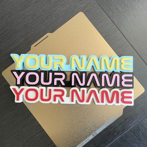 Pode incluir: Três placas de nome personalizadas empilhadas em uma superfície dourada. A placa superior é azul claro com letras amarelas, a placa do meio é preta com letras rosa e a placa inferior é branca com letras vermelhas. O texto em cada placa diz "YOUR NAME".