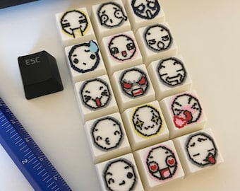May include: A collection of white square keycaps with various emoji faces drawn on them. The keycaps are arranged on a white surface, with a black ESC key and a blue ruler visible. The emoji designs include expressions of joy, sadness, and anger.