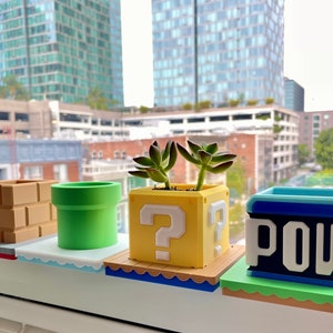 Puede incluir: Cuatro macetas decorativas en un alféizar, cada una con un tema de videojuego. El conjunto incluye un bloque de ladrillos, una tubería verde, un bloque amarillo con un signo de interrogación y un bloque POW azul. Cada maceta contiene una pequeña planta suculenta.