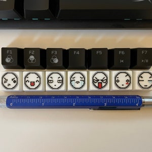 May include: A collection of black and white keyboard keycaps with various symbols and emoji faces. A blue and silver multi-tool pen with ruler markings is also present. The keycaps are arranged in rows, showcasing a unique design.