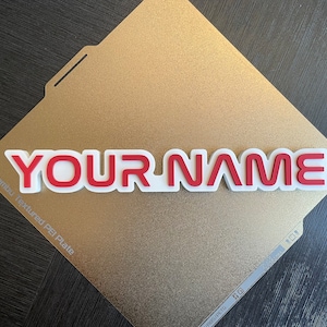Può includere: Una targhetta personalizzata in rosso e bianco, con la scritta "YOUR NAME", è esposta su una superficie dorata e strutturata. La targhetta è stampata in 3D. Lo sfondo è una piastra PEI strutturata dorata.